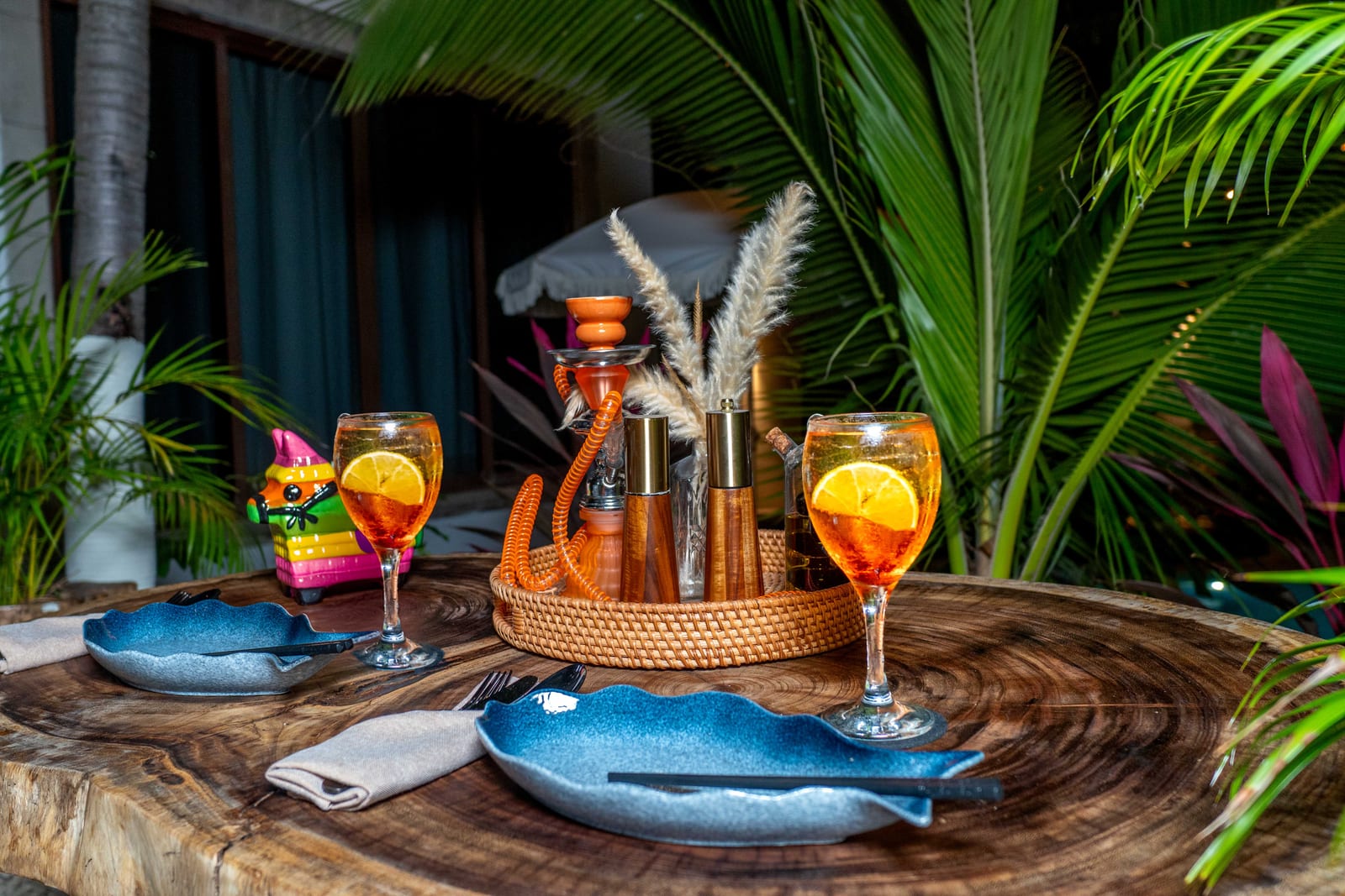 Mesa nocturna de MERO 18 entre palmas, con cocteles y platos azules en el patio selvático