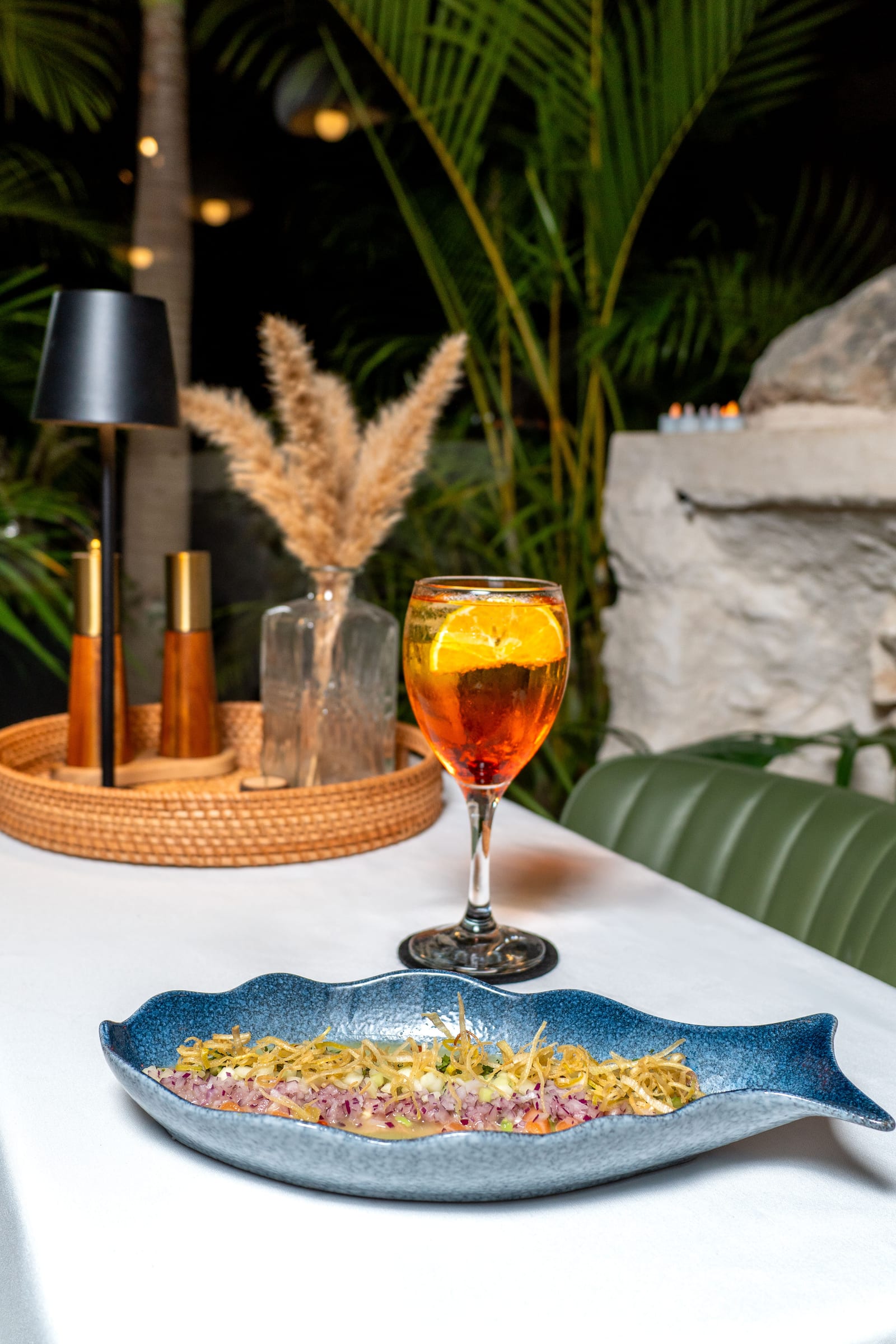 Ceviche en plato azul con forma de pez y aperol spritz sobre mesa nocturna entre palmas en MERO 18